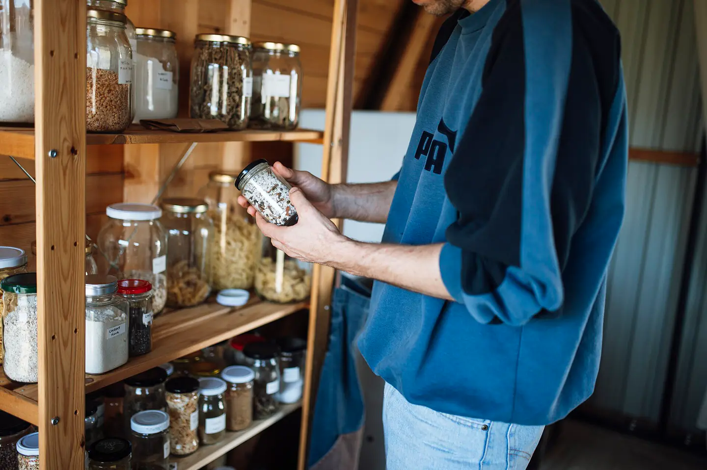 Person examines a jar of spice on a wooden shelf in a kitchen pantry during daytime in a cozy home setting