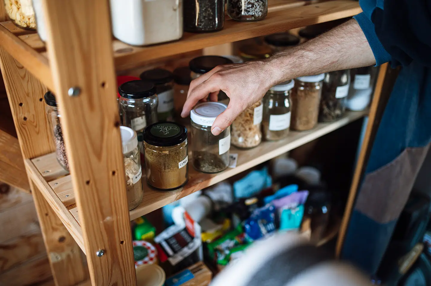 Person reaches for a jar on a wooden shelf filled with various containers in a kitchen or pantry setting